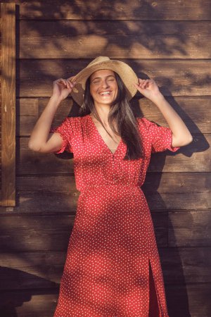 Foto de Mujer joven positiva en vestido de lunares rojos y sombrero sonriendo con los ojos cerrados mientras está de pie sobre fondo de madera en el día soleado - Imagen libre de derechos