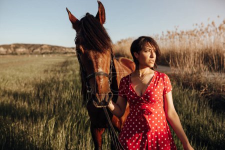 Foto de Joven hembra en vestido rojo largo con caballo mirando hacia otro lado mientras está de pie en el patio del rancho en el día soleado en el campo - Imagen libre de derechos