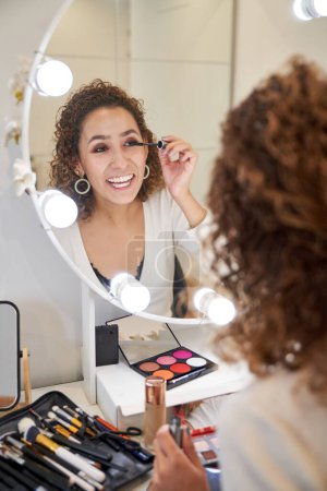 Téléchargez les photos : Esthéticienne souriante mettant un mascara sur les cils tout en se maquillant les yeux en regardant dans un miroir avec des lampes éclairées - en image libre de droit