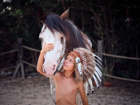 Téléchargez les photos : Petit garçon à l'intérieur du paddock avec bonnet de guerre sur la tête debout devant un magnifique cheval et le caresser - en image libre de droit