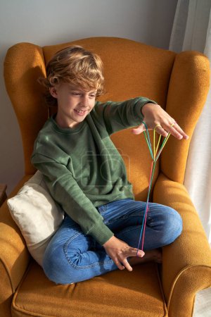 Foto de Alto ángulo de niño preadolescente positivo en ropa casual descansando en un sillón y disfrutando de un juguete de cuerda mientras hace diferentes combinaciones en casa - Imagen libre de derechos