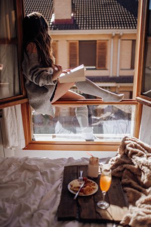 Foto de Mujer joven sentada con sabrosa bebida y libro en la ventana - Imagen libre de derechos