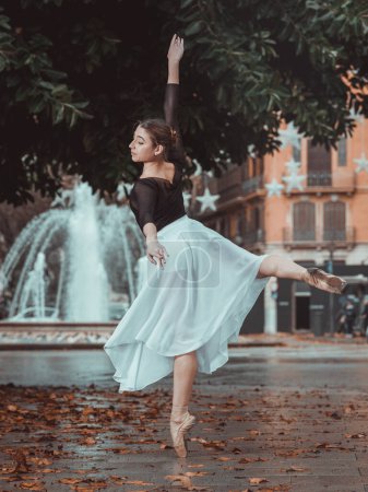 Foto de Vista lateral de la fina y elegante bailarina de ballet femenina en zapatos puntiagudos y tutú de seda realizando posición arabesca en el parque cerca de la fuente - Imagen libre de derechos