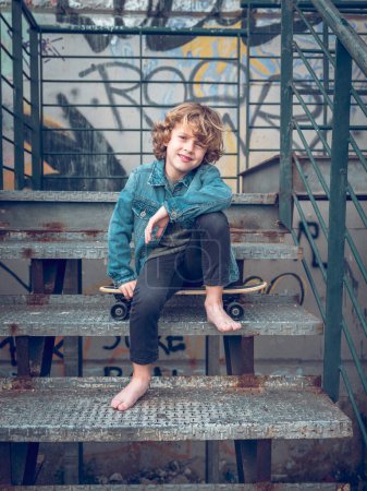 Photo for Full body of confident boy in casual clothes sitting on penny board on shabby steps while smiling and looking at camera - Royalty Free Image