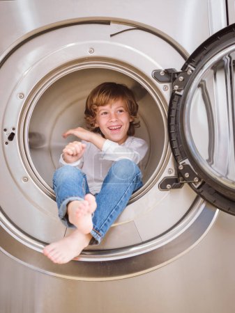 Foto de Cuerpo completo de niño descalzo encantado mirando hacia otro lado mientras está sentado en el tambor de la lavadora moderna en la sala de lavandería de luz - Imagen libre de derechos