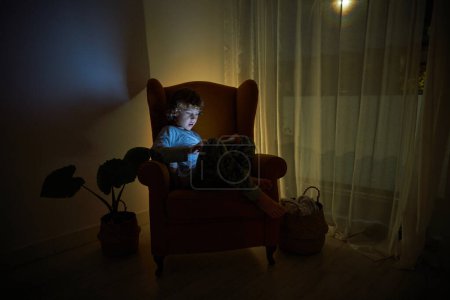 Photo for Focused boy playing game on tablet while sitting in comfortable armchair in dark room with flowers in evening at home - Royalty Free Image