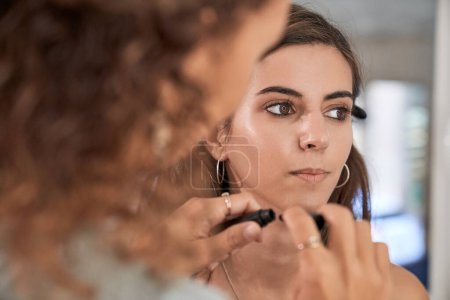 Téléchargez les photos : Crop maquilleuse anonyme appliquant un produit cosmétique sur un modèle aux cheveux foncés assis dans un studio de beauté clair sur fond flou - en image libre de droit