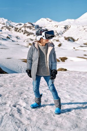 Téléchargez les photos : Corps entier d'un enfant anonyme portant une veste chaude et un casque VR debout sur la neige près des collines enneigées pendant le jeu de la journée d'hiver - en image libre de droit