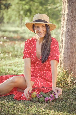 Téléchargez les photos : Jeune femme en robe d'été et chapeau de paille avec assis sur l'herbe dans le parc - en image libre de droit