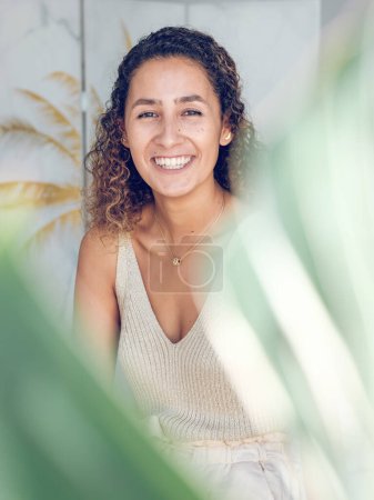 Téléchargez les photos : Femme gaie avec des cheveux bouclés bruns dans des vêtements décontractés regardant caméra contre mur clair derrière la plante verte - en image libre de droit