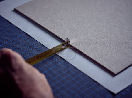 Photo for Unrecognizable crop male artisan using tape and measuring paper on blue mat while working in bookbinding workshop - Royalty Free Image