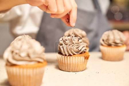 Téléchargez les photos : Concentration sélective de la culture cuisinier méconnaissable dans tablier saupoudrer cupcakes maison avec des miettes de chocolat dans la cuisine - en image libre de droit