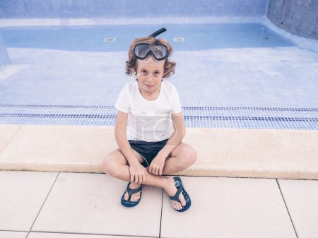 Foto de Desde arriba de un niño de cuerpo completo con conjunto de natación y sentado con las piernas cruzadas mientras descansa en el borde de la piscina - Imagen libre de derechos