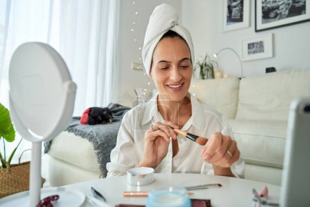 Photo for Cheerful adult female blogger with towel on head applying foundation on hand with cosmetic brush while recording beauty blog on smartphone and looking down - Royalty Free Image