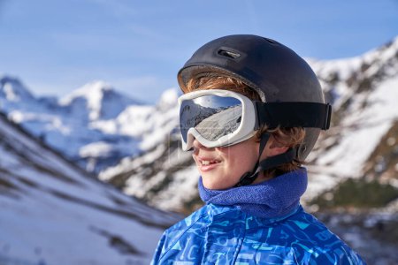 Téléchargez les photos : Enfant positif en lunettes de protection et casque souriant et regardant loin sur fond flou de montagnes enneigées par une journée ensoleillée sur la station de ski - en image libre de droit