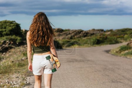 Téléchargez les photos : Vue arrière du jeune skateboarder féminin méconnaissable avec longboard se promenant sur la passerelle contre les montagnes et la mer - en image libre de droit