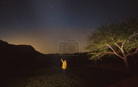 Téléchargez les photos : Pleine longueur d'enfant en veste jaune sur l'herbe près de la montagne et des arbres admirant le ciel étoilé - en image libre de droit