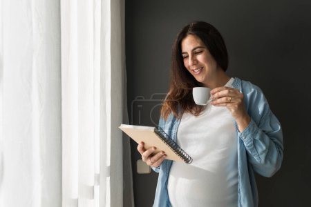Photo for Cheerful pregnant female with cup of drink and notepad standing in room at home - Royalty Free Image