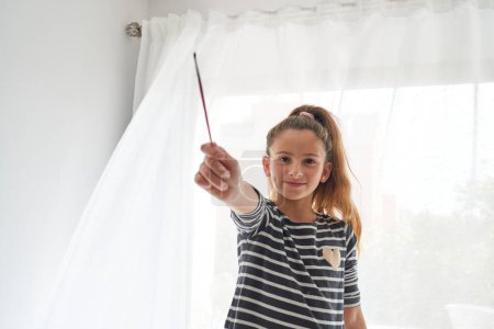 Photo for Positive child in striped long sleeve demonstrating paintbrush standing near window with white curtain and looking at camera on sunny morning - Royalty Free Image