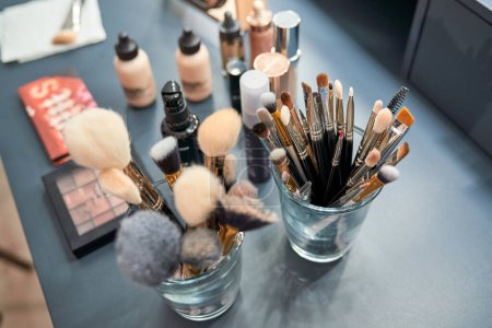 Téléchargez les photos : De la collection ci-dessus de divers pinceaux cosmétiques dans des verres placés sur la table avec des produits de maquillage assortis dans un salon de beauté moderne - en image libre de droit