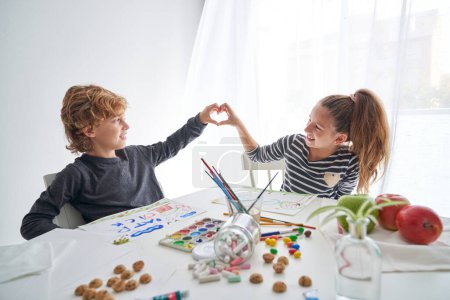 Téléchargez les photos : Enfants positifs se regardant et faisant un geste en forme de cœur assis à table avec des peintures dans un studio de lumière - en image libre de droit