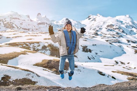 Photo for Happy kid wearing warm clothes and hat with mittens jumping high with raised arms near snowy hills under blue sky and looking away in sunny winter day - Royalty Free Image