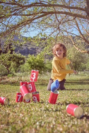 Foto de Cuerpo completo de niño pequeño jugando con coloridas latas derribadas mientras pasa tiempo en el parque - Imagen libre de derechos