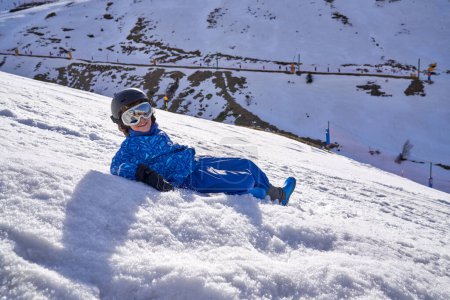 Téléchargez les photos : Garçon positif en vêtements de dessus avec lunettes et casque souriant et couché sur la pente de montagne recouvert de neige blanche par une journée ensoleillée sur la station de ski - en image libre de droit