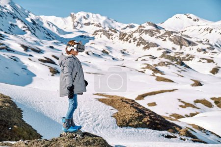 Foto de Vista lateral de cuerpo completo de un niño irreconocible que usa ropa exterior y auriculares VR explorando el mundo virtual mientras está parado en suelo rocoso cerca de montañas nevadas - Imagen libre de derechos