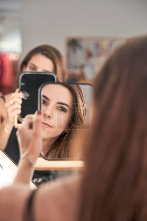 Téléchargez les photos : Cultiver modèle féminin attrayant avec les cheveux longs regardant miroir à la main tout en étant assis dans un salon de beauté moderne léger pendant le rendez-vous - en image libre de droit