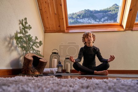 Photo for Full body of barefoot boy meditating with closed eyes while sitting in Padmasana on yoga mat at home - Royalty Free Image