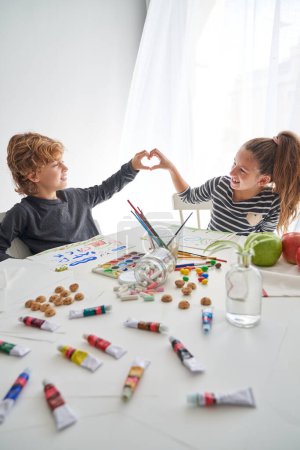 Téléchargez les photos : Des enfants optimistes faisant des gestes cardiaques et se regardant tout en s'asseyant à table avec diverses peintures et dessins colorés - en image libre de droit