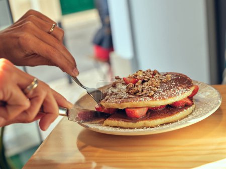 Téléchargez les photos : Cultiver une personne anonyme coupant de savoureuses crêpes sucrées avec de la pâte de chocolat et des fraises tout en étant assis à table dans un café pendant le petit déjeuner - en image libre de droit