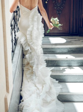 Téléchargez les photos : Vue arrière de la culture mariée anonyme en robe de mariée blanche élégante avec volants et dentelle marchant sur les marches avec bouquet de fleurs à l'entrée de l'hôtel - en image libre de droit