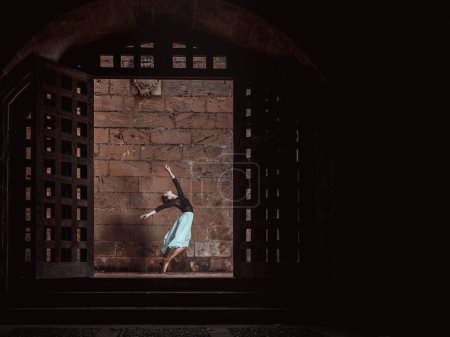 Photo for Side view of graceful slim ballerina dancing in dark hall near brick wall and gates - Royalty Free Image