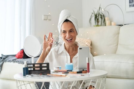Téléchargez les photos : Heureuse blogueuse adulte avec serviette sur la tête regardant miroir avec friseur de cils tout en faisant maquillage et enregistrement blog de beauté sur smartphone à la maison le matin - en image libre de droit
