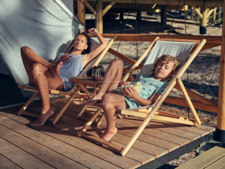 Foto de Cuerpo completo de madre con hijo sentado en salones mientras descansan juntos en la terraza de madera en el día de verano en el campo - Imagen libre de derechos