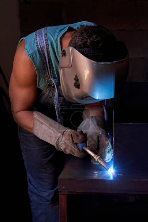 Téléchargez les photos : Masculin sans visage dans un masque de protection utilisant une machine à souder pour souder la structure métallique tout en travaillant dans un atelier sombre avec une lumière tamisée - en image libre de droit