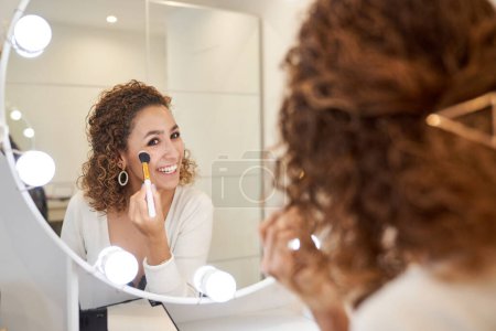 Photo for Cheerful young woman doing facial makeup of powder with brush looking at mirror in dressing room - Royalty Free Image