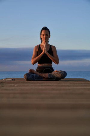 Photo for Full body of calm Hispanic female with closed eyes practicing lotus pose with namaste hands during yoga session on shore - Royalty Free Image