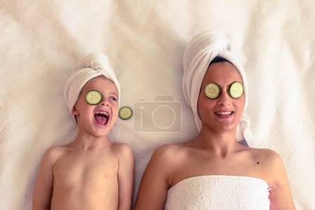 Photo for From above of happy woman and little son relaxing on white blanket with towels on heads while having skincare session with cucumber slices on eyes - Royalty Free Image