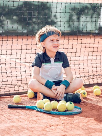 Photo for Full length of content little boy with blond hair in activewear smiling and looking away while resting on tennis court with crossed legs near scattered balls and racket after match - Royalty Free Image