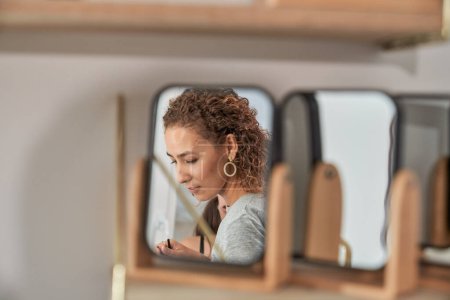 Foto de Mujer con el pelo rizado reflejándose en un pequeño espejo en la pared mientras está de pie con productos cosméticos en el estudio de luz durante la jornada laboral - Imagen libre de derechos