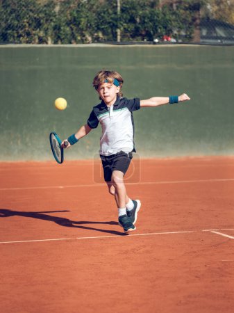 Téléchargez les photos : Corps complet de petit garçon actif en vêtements de sport et bandeau jouant au tennis avec raquette sur le terrain par une journée ensoleillée - en image libre de droit