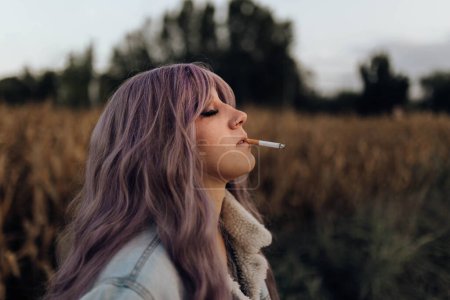 Foto de Vista lateral de la hermosa mujer moderna con el pelo rosa fumar cigarrillo con los ojos cerrados en la naturaleza otoñal - Imagen libre de derechos
