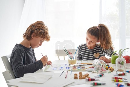 Téléchargez les photos : Des enfants concentrés dessinent des images à l'aide de pinceaux et de peinture aquarelle assis à table avec divers bonbons dessus - en image libre de droit