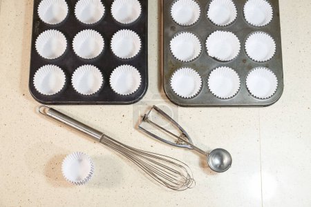 Photo for From above of metal cupcake baking trays with liners placed on table with whisk and scoop spoon in light kitchen - Royalty Free Image