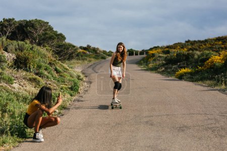 Téléchargez les photos : Cropped méconnaissable ethnique navigation féminine sur smartphone tout en étant assis longboard sur route asphaltée - en image libre de droit