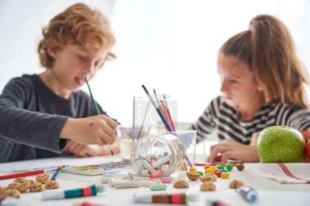 Téléchargez les photos : Enfants focalisés peignant avec des pinceaux assis à une table désordonnée avec des fournitures d'art dispersées et des biscuits sucrés dans la pièce lumineuse - en image libre de droit