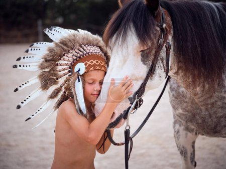 Foto de Vista lateral de un niño lindo con los ojos cerrados con ropa de cabeza india tradicional con plumas acariciando hocico de caballo gris en el paddock contra el fondo borroso - Imagen libre de derechos